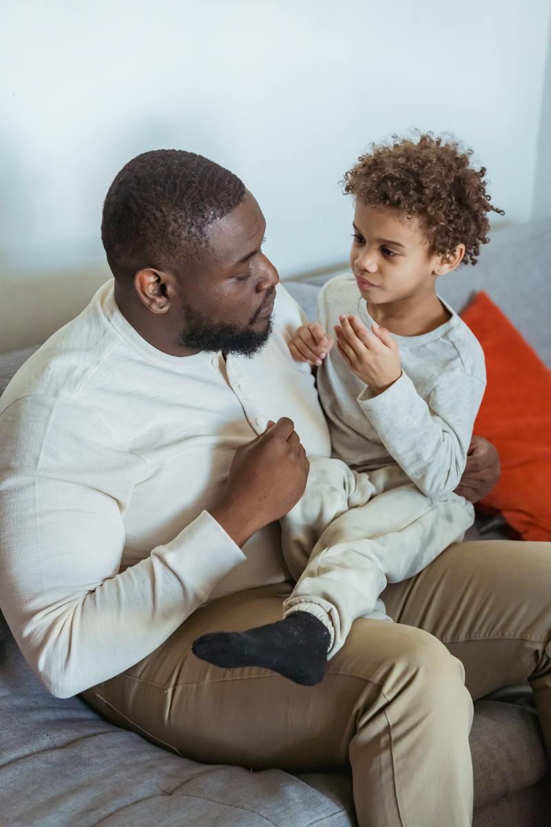 From above of African American father holding little child and having serious conversation while sitting on couch at home